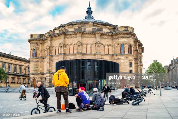 Young adults play with bikes and skateboards on Bristo Square with McEwan Hall of the University of Edinburgh in the background, in downtown Edinburgh, Scotland, UK on an overcast day.